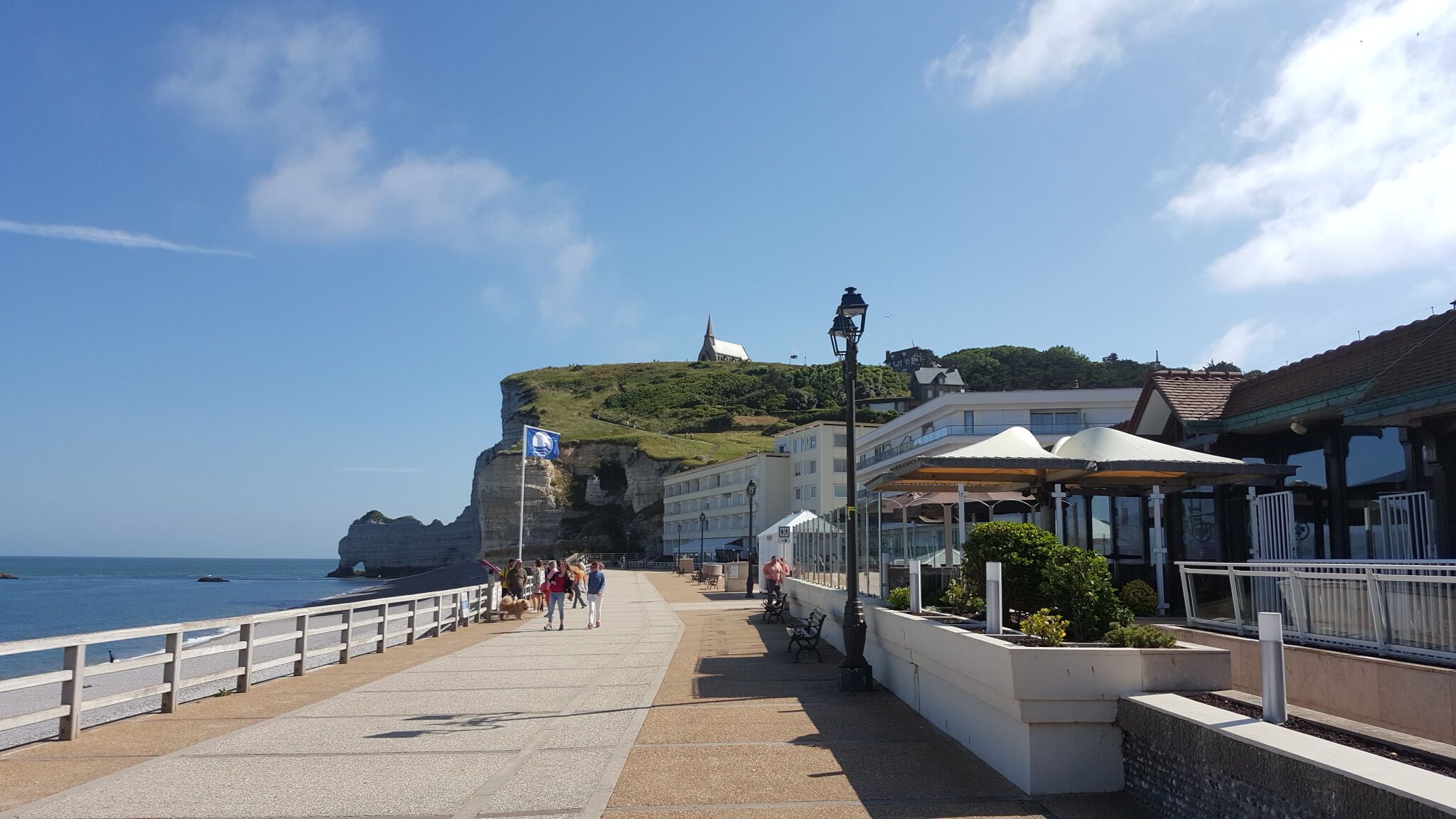 A coastal promenade with a rocky cliff, a church on top, and a row of buildings with outdoor seating.