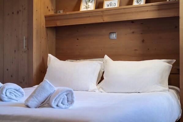 A neatly made bed with white linens and two pillows, a wooden headboard, and a shelf above with framed pictures and a sign.