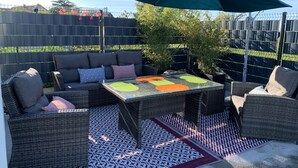 En udendørs patio med fletmøbler, et glasbord med farverige bordskåner og en stor parasol.