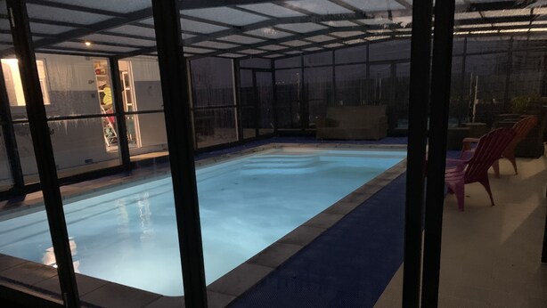 En indendørs swimmingpool med et glasomhegning, omgivet af et mønstret gulv og et par stole.