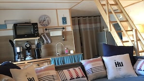 Interior acolhedor de uma cabana com teto de madeira, uma kitchenette com micro-ondas, cafeteira e pia, um sofá com almofadas de temática náutica e uma escada que leva a um andar superior.