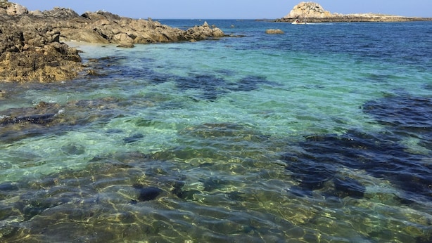 Cena costeira com águas turquesa cristalinas, formações rochosas e uma ilha ao longe.