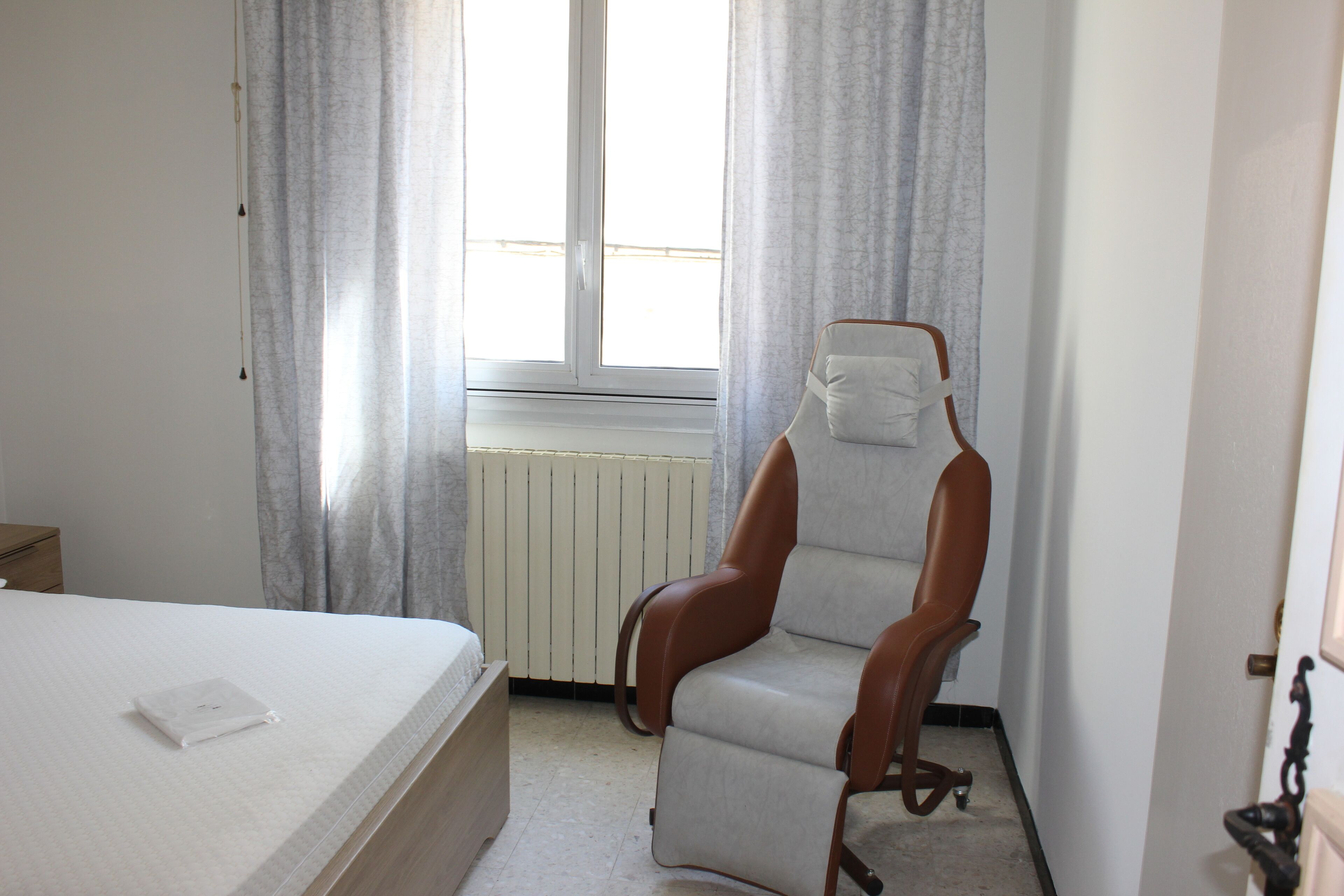 A hotel room with a bed, a chair, a radiator, and a window with curtains.