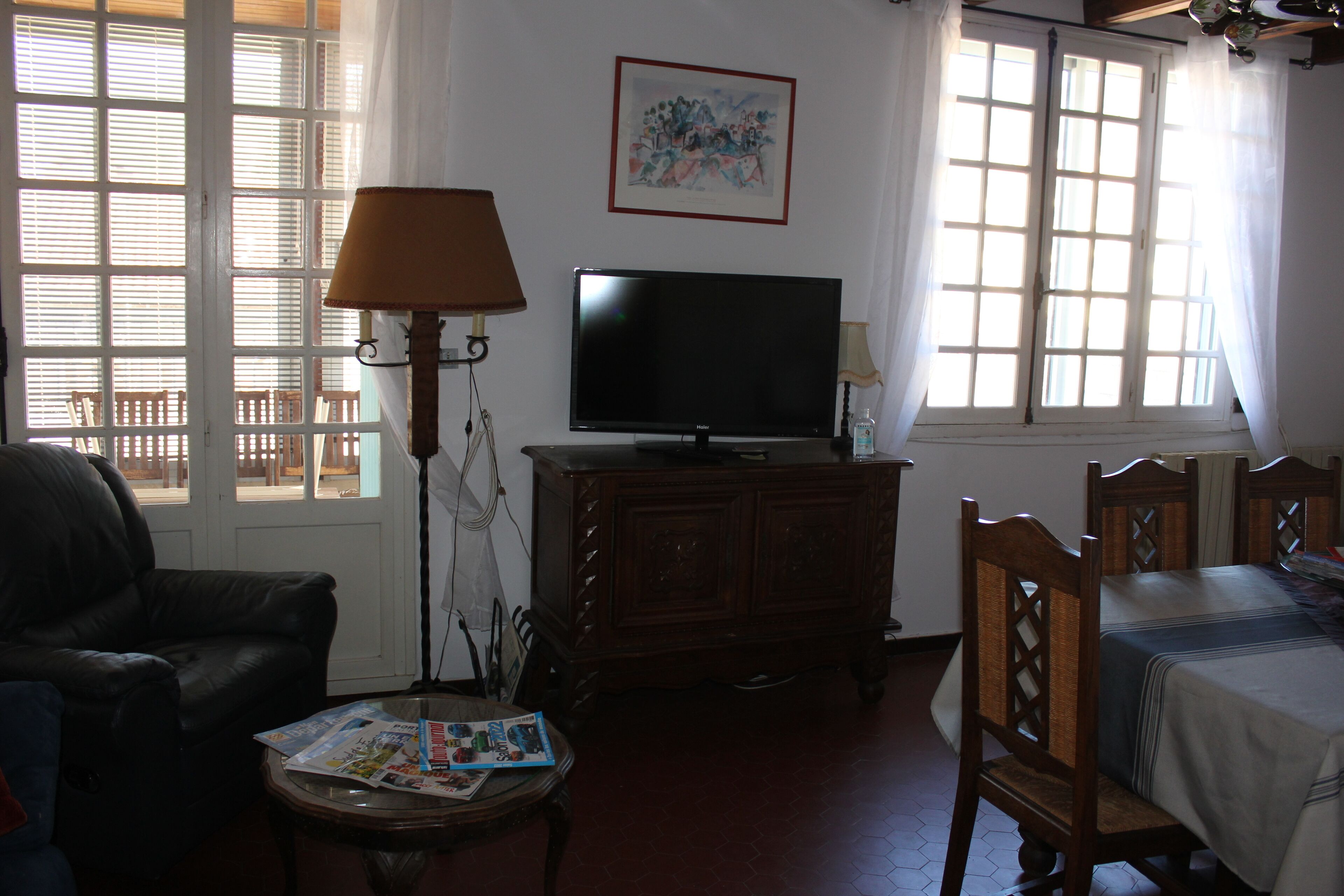 Una sala de estar con televisión sobre un mueble de madera, un sofá, una mesa de centro con revistas y una cama con cubrecama blanco.