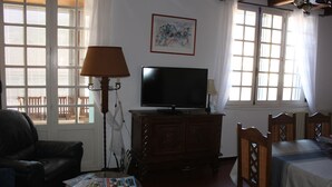 Una sala de estar con televisión sobre un mueble de madera, un sofá, una mesa de centro con revistas y una cama con cubrecama blanco.