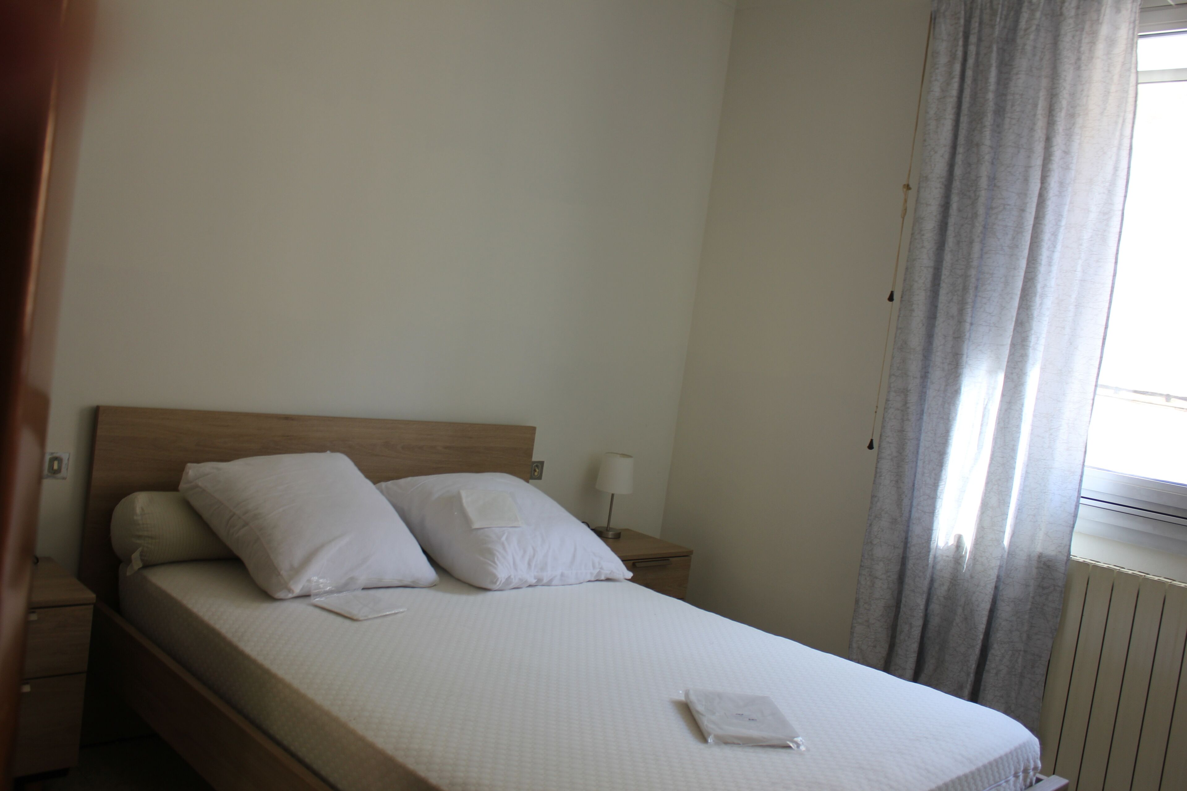 Een net opgemaakt bed met witte lakens en kussens, een houten hoofdbord, een nachtkastje met een lamp, en een raam met gordijnen.