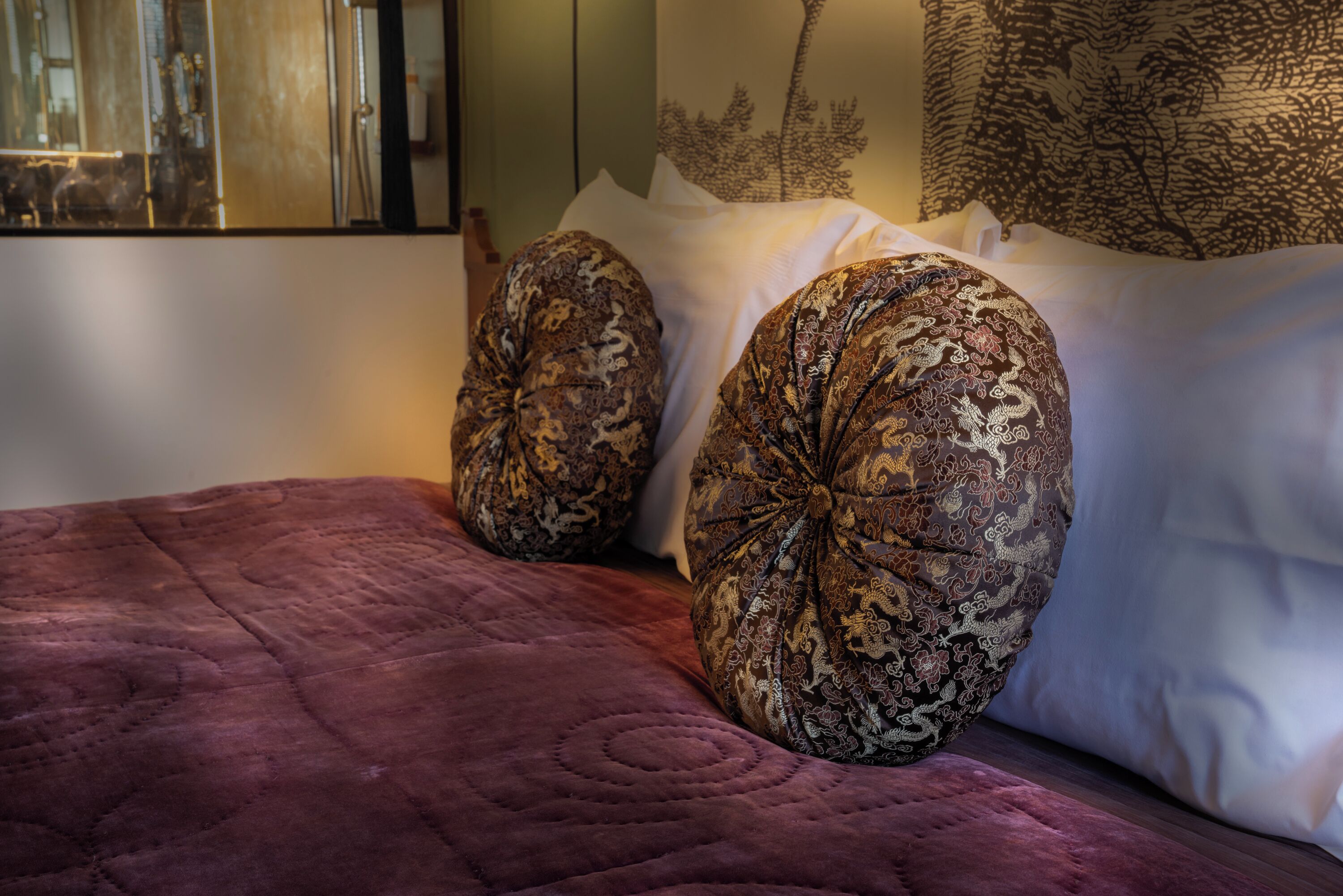Een bed met een paars quilt, twee gestreepte kussens en een muur met bladmotief.