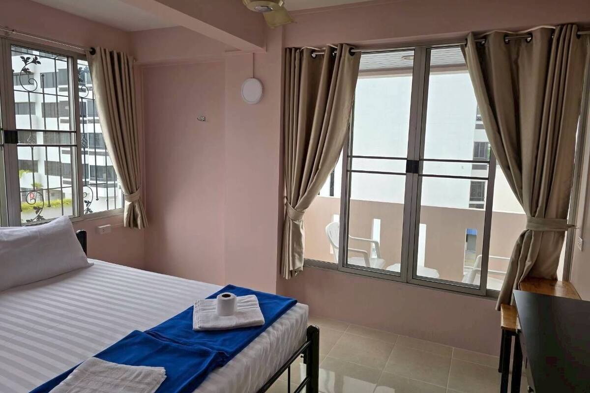 A hotel room with a bed, a ceiling fan, a window with curtains, and a view of the outside.