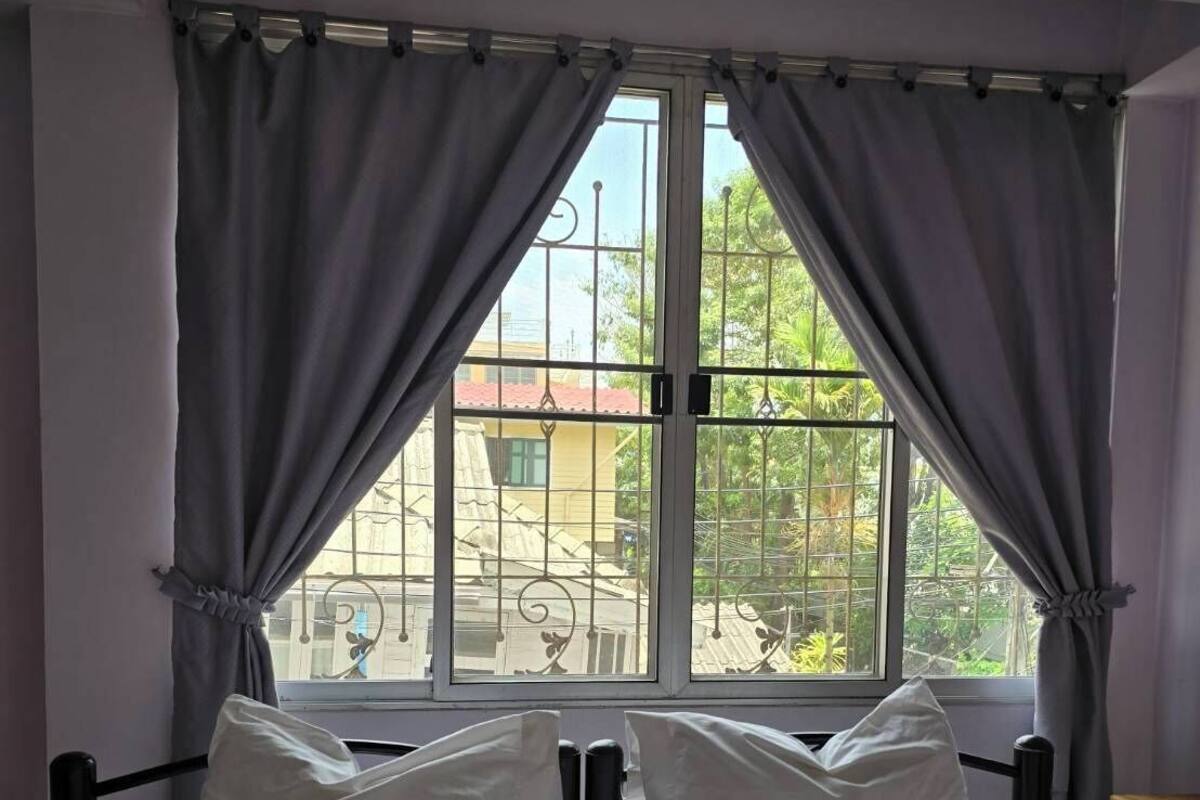 A bed with white linens and two pillows, a window with metal bars, and grey curtains.