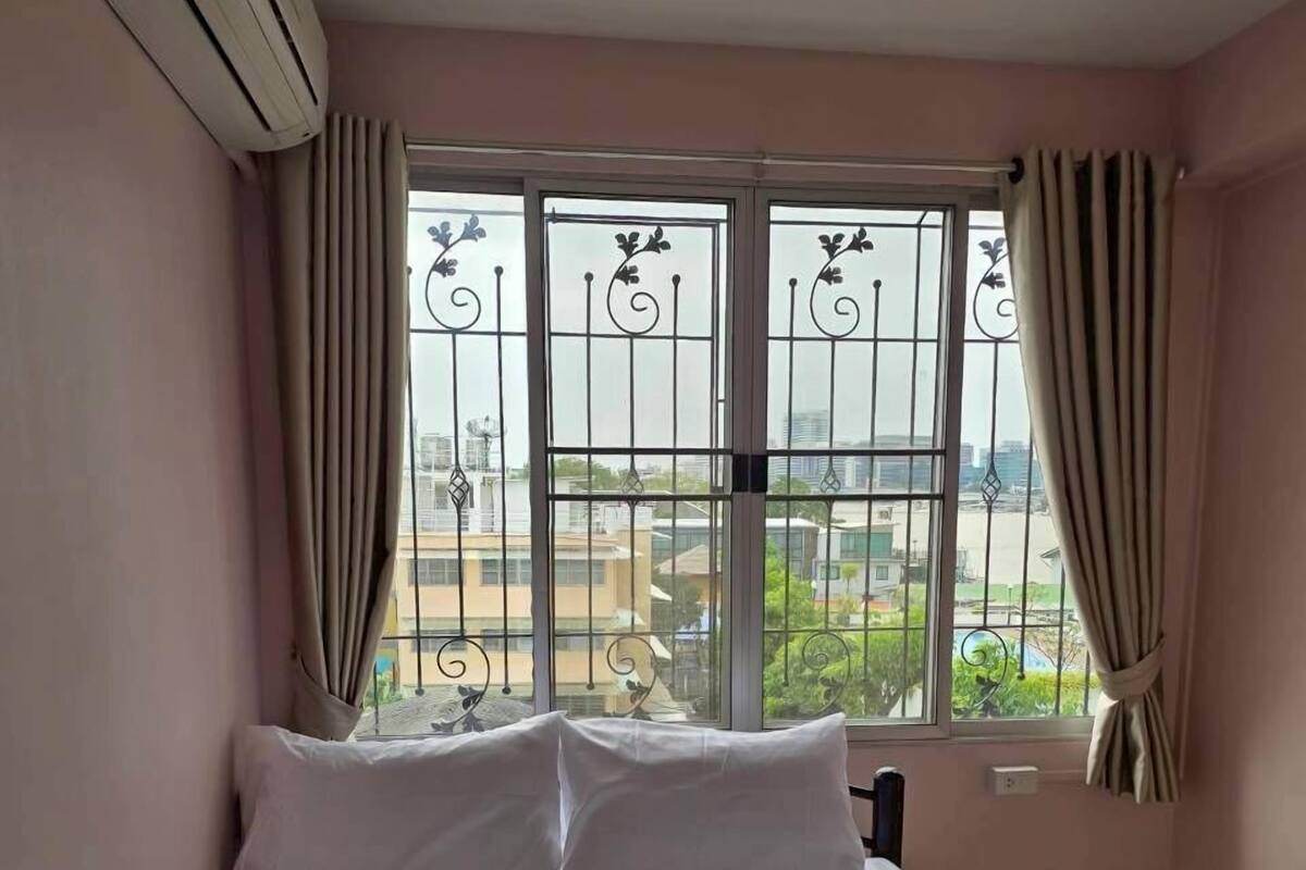 A single-bed room with a striped bedspread, two pillows, a window with metal bars, and curtains.