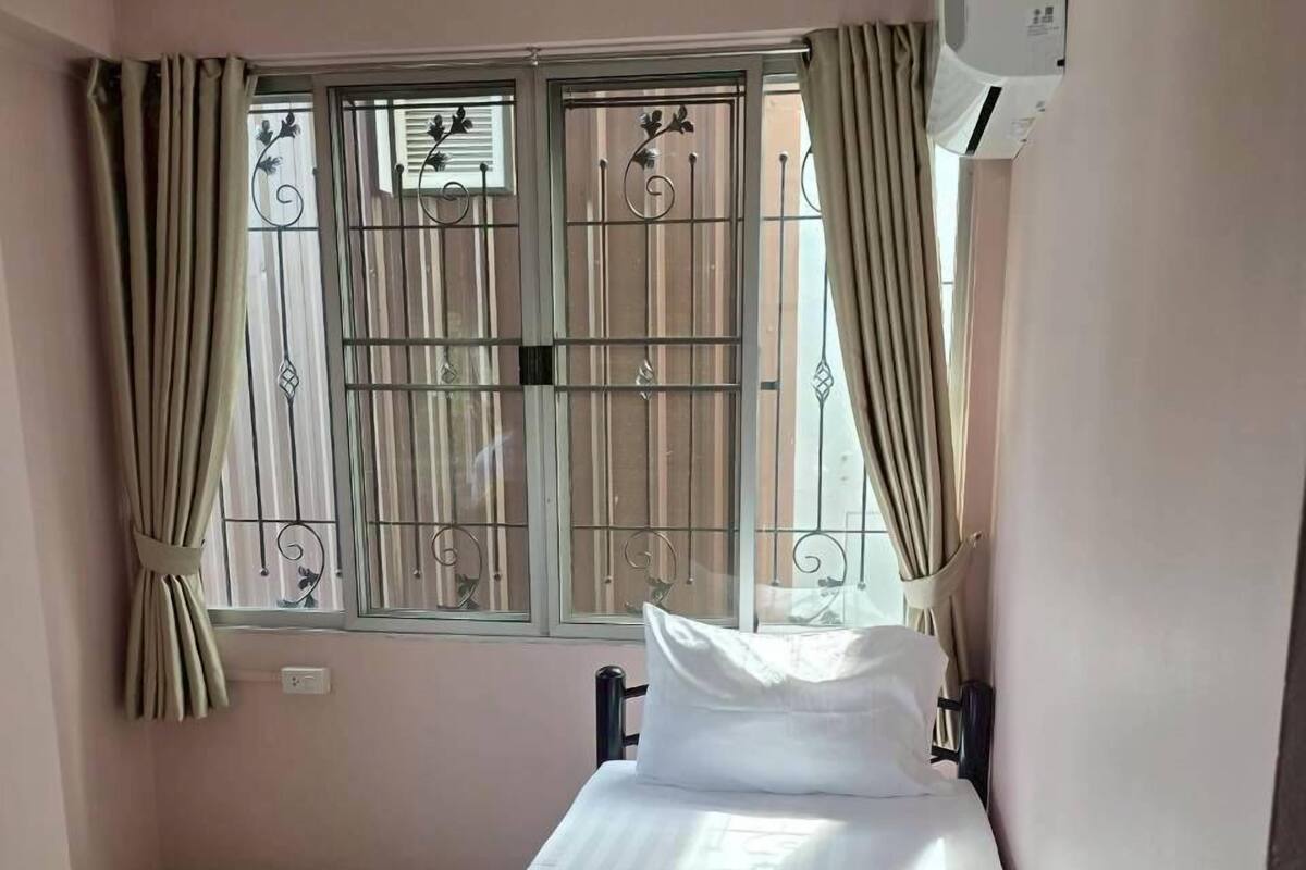 A single bed with a blue blanket and a roll of toilet paper, a window with metal bars, and beige curtains.