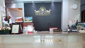 A hotel reception desk with a black backdrop displaying 'The Hotel Beauty' logo, framed certificates, brochures, and a 'Check In' sign. - The Beauty Hotel (Gangneung)