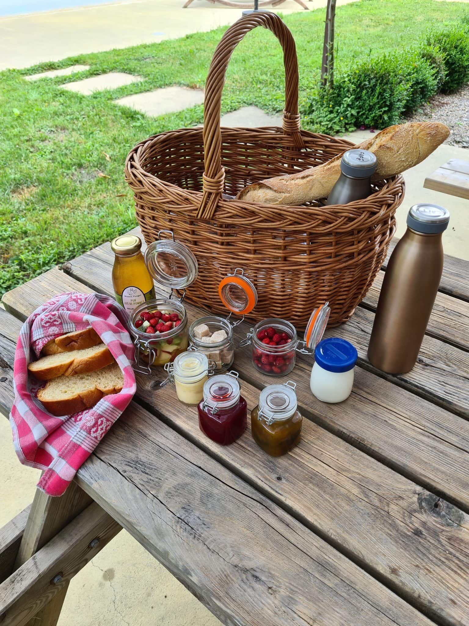 Een picknickopstelling op een houten tafel met een rieten mand, brood, potten jam en een waterfles.