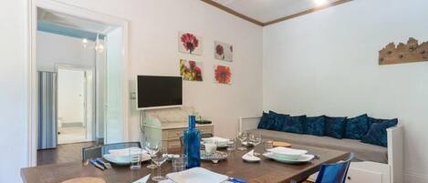 A dining area with a wooden table set for four, a sofa with blue cushions, and a television on the wall.