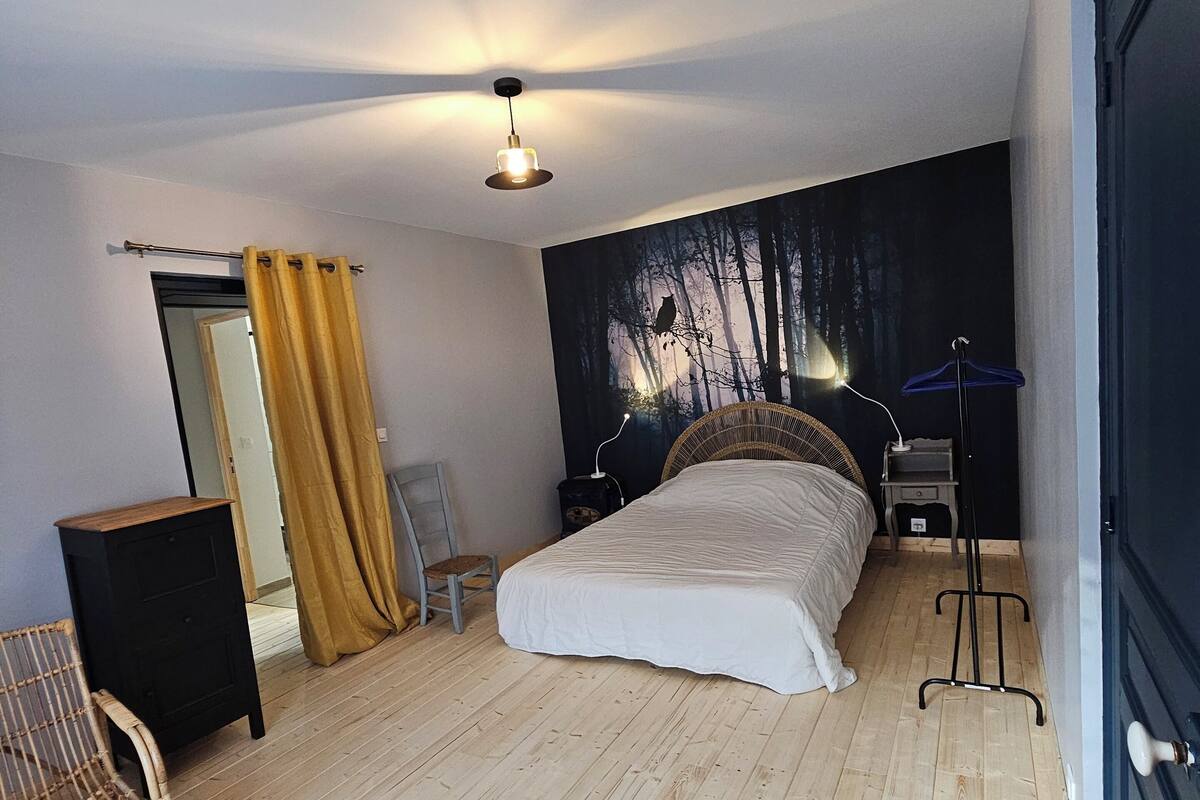 A bedroom with a bed, a chair, a small table, a wardrobe, and a wall with a forest mural.