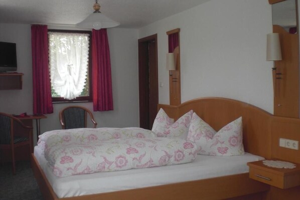A bedroom with a wooden bed, floral bedding, a desk with a chair, a window with curtains, and a mirror on the wall. - Gasthaus Pension Donishäusle (Titisee-Neustadt)