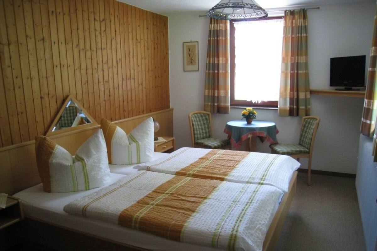 Hotelkamer met houten hoofdeinde, bed met wit en gestreept beddengoed, een klein rond tafeltje met een bloemenvaas, twee stoelen en een televisie op een standaard.