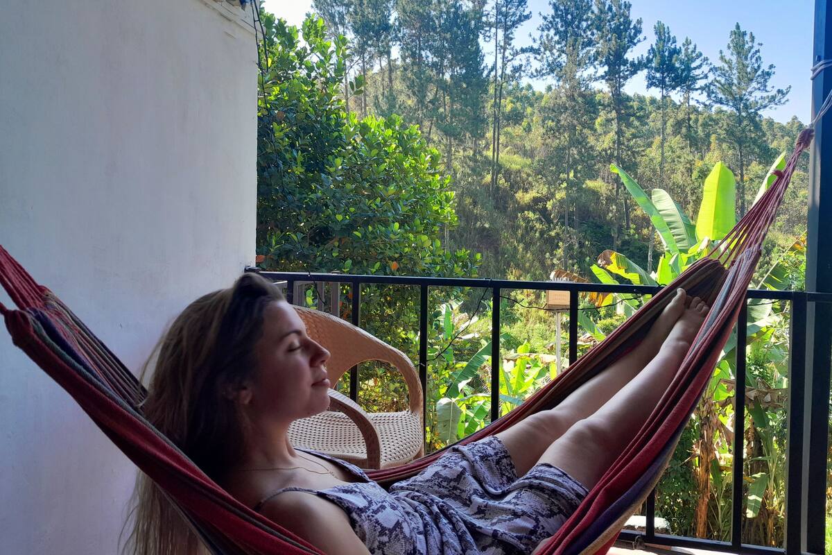 A person relaxing in a hammock on a balcony with a view of lush greenery and trees.