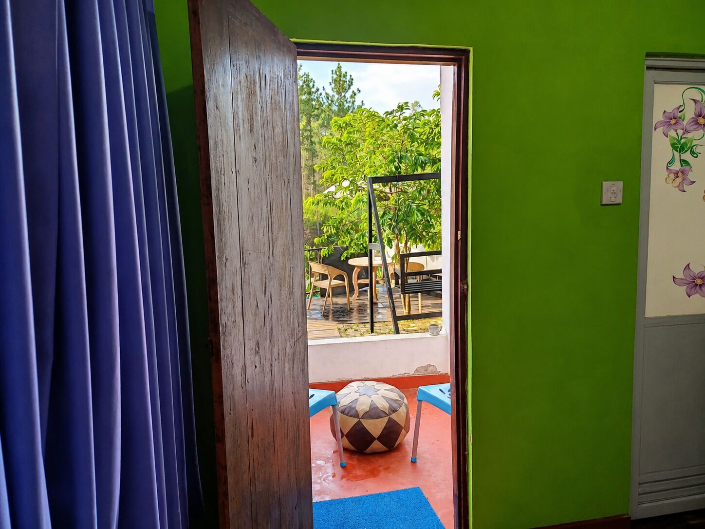 Una habitación con puerta de madera, cortina azul, pared verde, y un pequeño espacio exterior con mesa y sillas, además de una pelota de colores en el suelo.