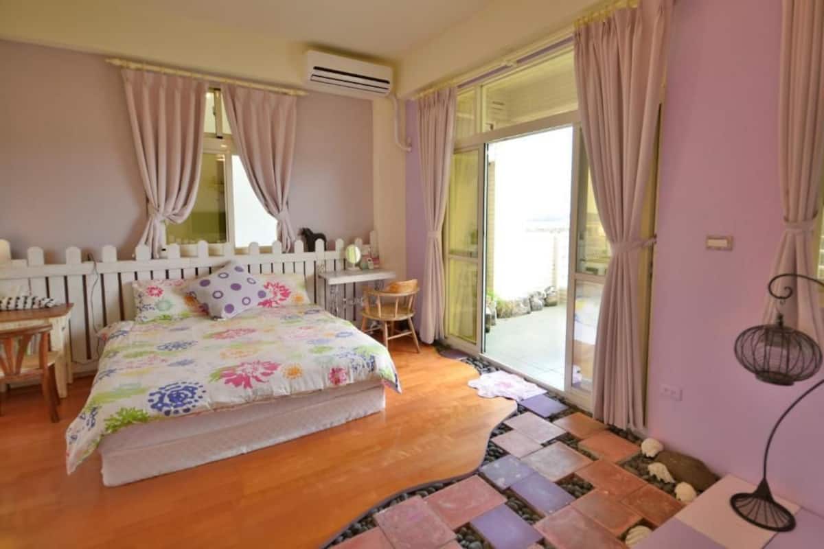 Quarto com cama, uma mesinha, uma cadeira, janela com cortinas e vista para o exterior.