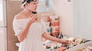 Una persona con vestido y sombrero blancos sostiene una taza, de pie en una cocina con un dispensador de agua y varios productos envasados en estantes.