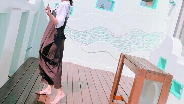 Una persona está de pie en una terraza de madera con una pared blanca ondulada y detalles en color turquesa al fondo.