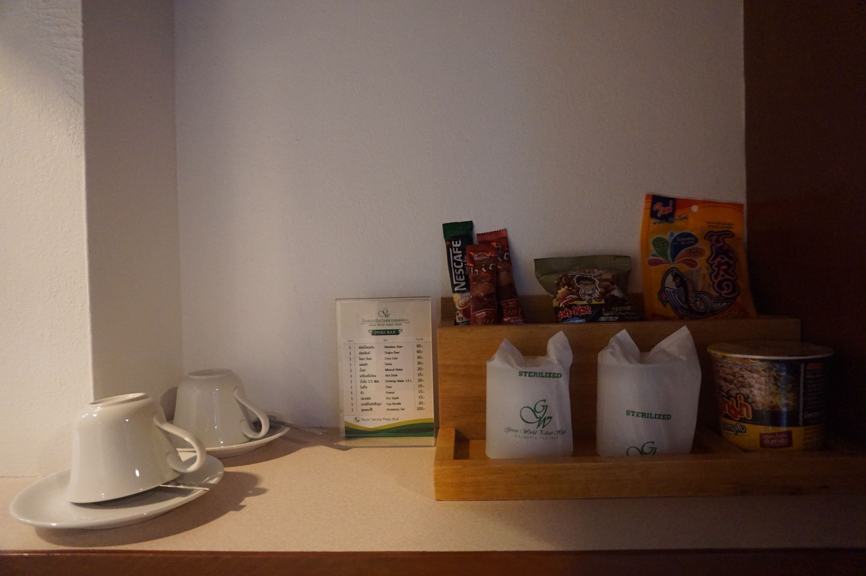 A breakfast setup with white cups and saucers, a menu, and a wooden tray with packaged food items and a can.