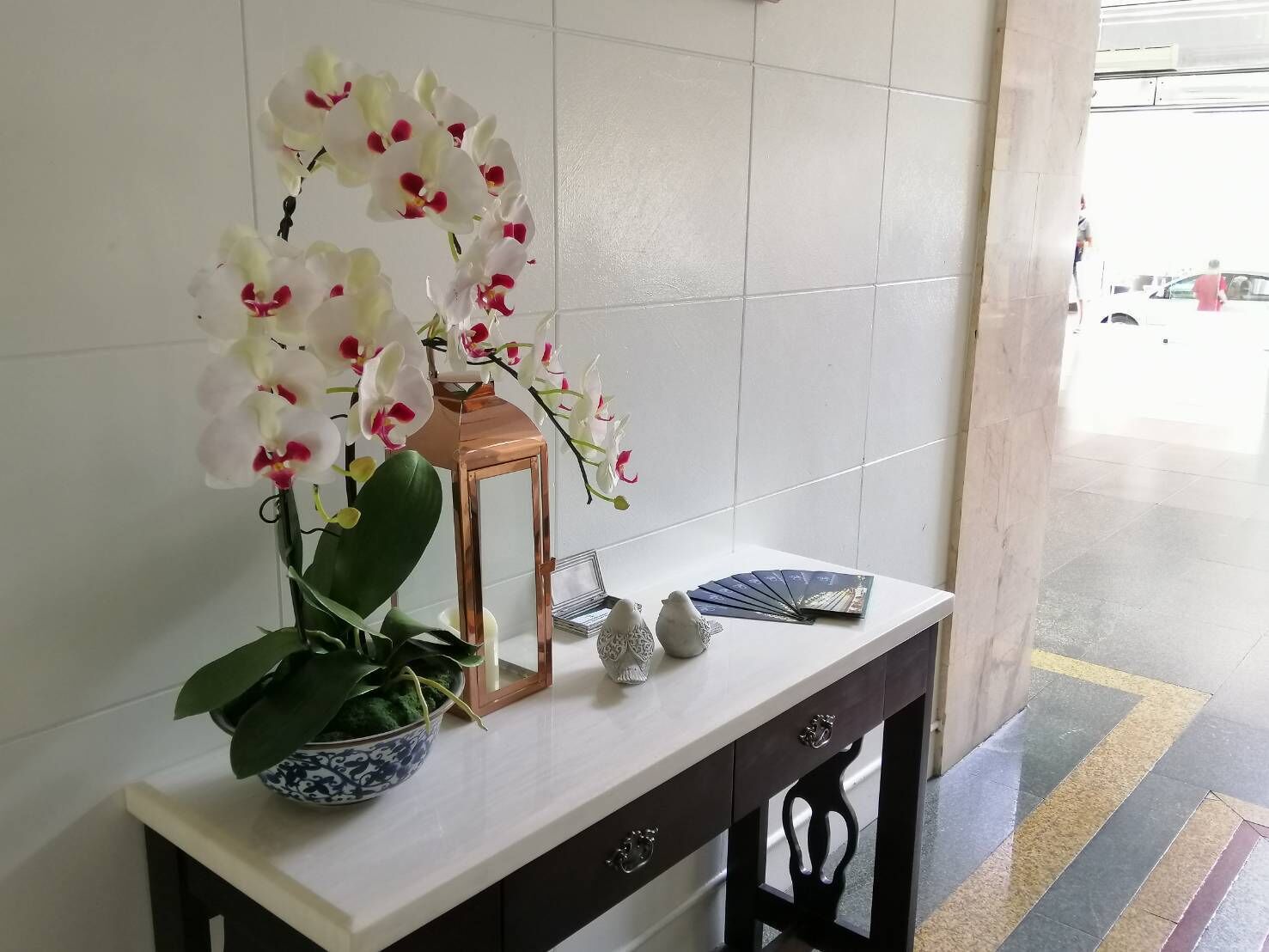 Una pared de azulejos blancos, una mesa blanca con un florero de orquídeas blancas, un farol pequeño y un abanico.