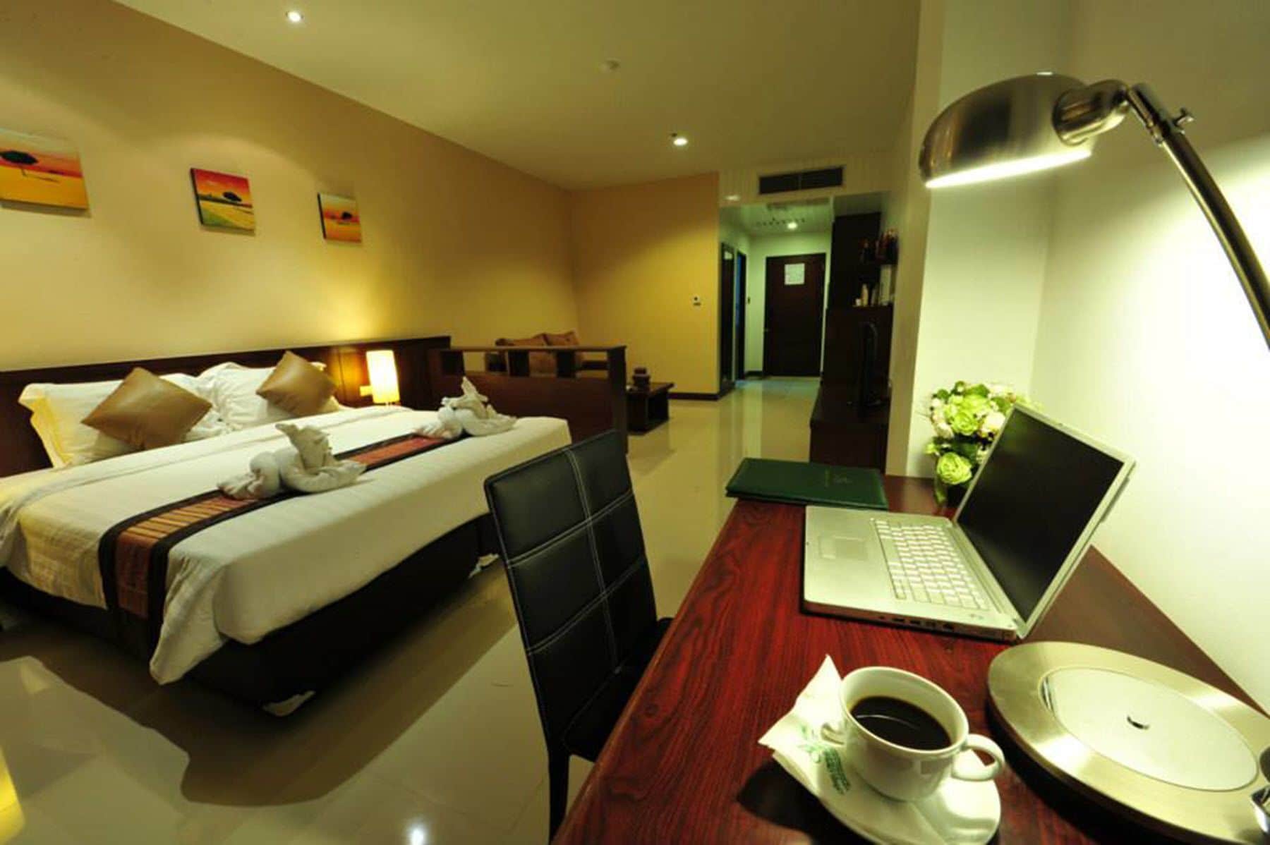 A hotel room with a bed, a desk with a laptop, a cup of coffee, and a vase of flowers.