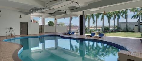 A swimming pool with a curved edge, surrounded by lounge chairs and a patio area with a view of palm trees and buildings.