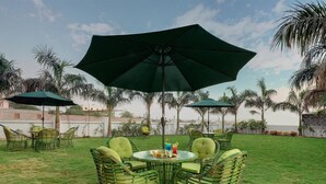 An outdoor seating area with wicker chairs and a table under a large umbrella, surrounded by palm trees and a clear sky.