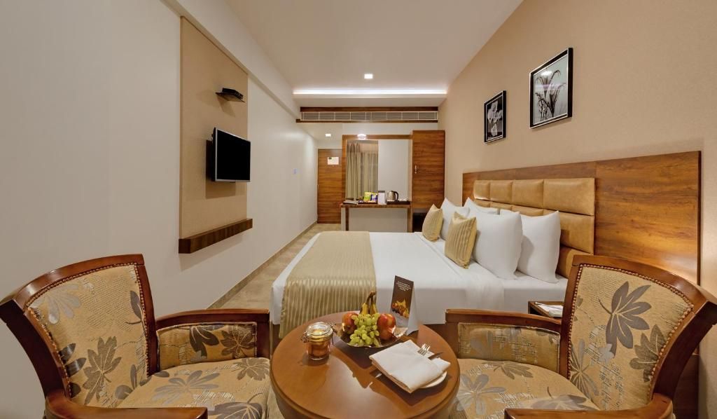 A hotel room with a bed, two armchairs, a small table with fruit, a television, and a mirror.