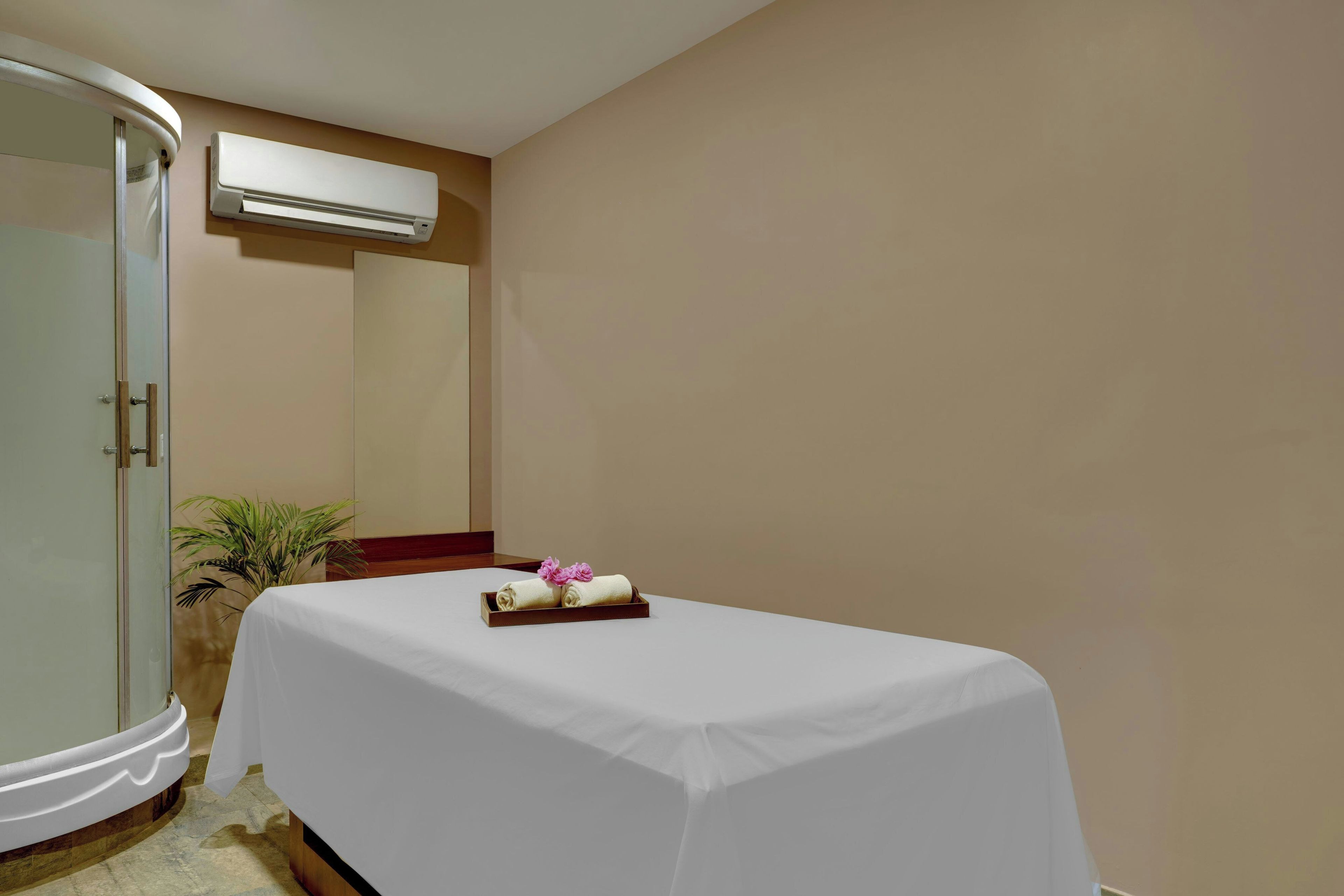 A hotel room with a bed covered in white, a small table with towels and flowers, a glass shower enclosure, and a wall-mounted air conditioner.