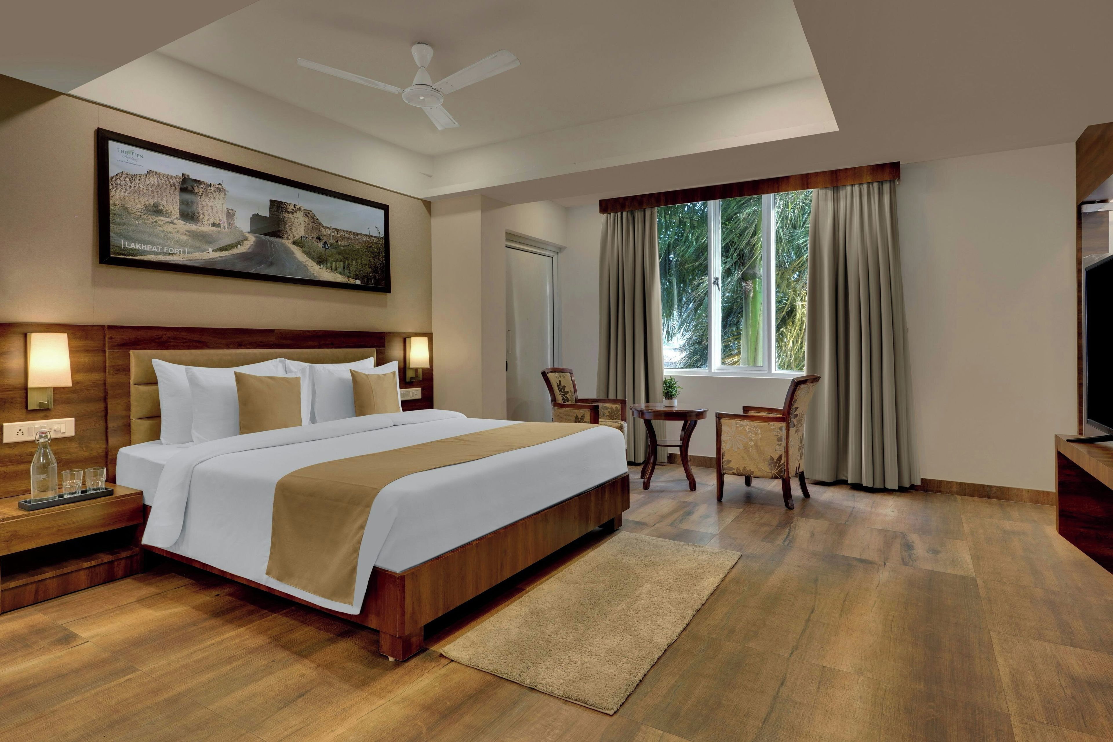 A hotel room with a large bed, a desk with a chair, a ceiling fan, a window with curtains, and a framed picture on the wall.