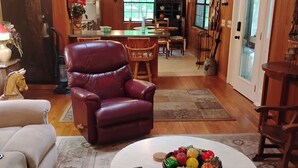 A cozy living room with a leather chair, a round coffee table with fruit, and a dining area in the background.
