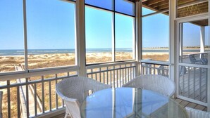 A covered outdoor area with a glass table and wicker chairs, overlooking a beach with dunes.