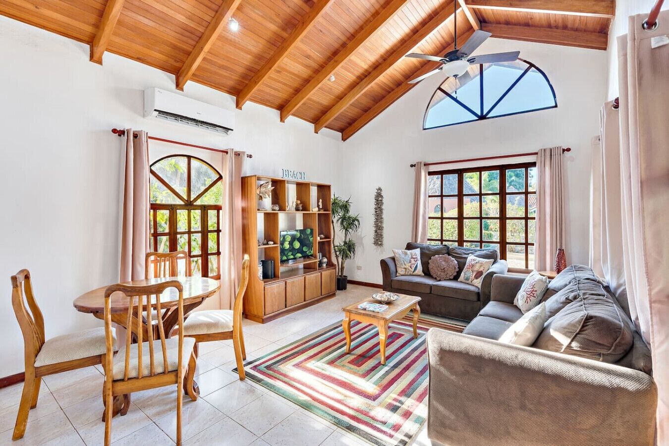 A spacious living room with wooden ceiling beams, a TV on a wooden stand, a sofa, and a dining table with chairs.