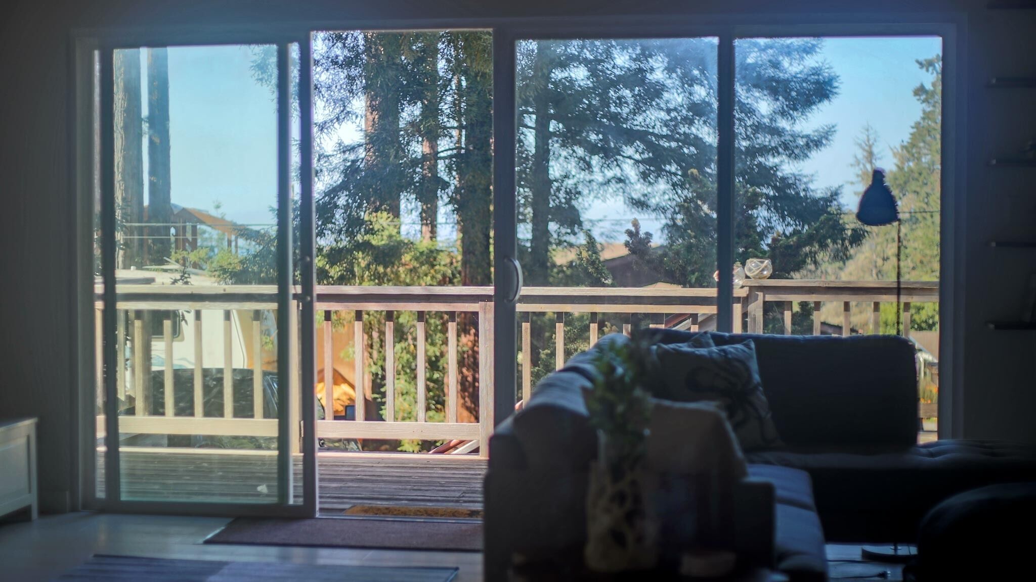 A room with a sliding glass door leading to a deck with a wooden railing, a sofa with cushions, and a small table with a vase.