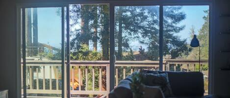 A room with a sliding glass door leading to a deck with a wooden railing, a sofa with cushions, and a small table with a vase.
