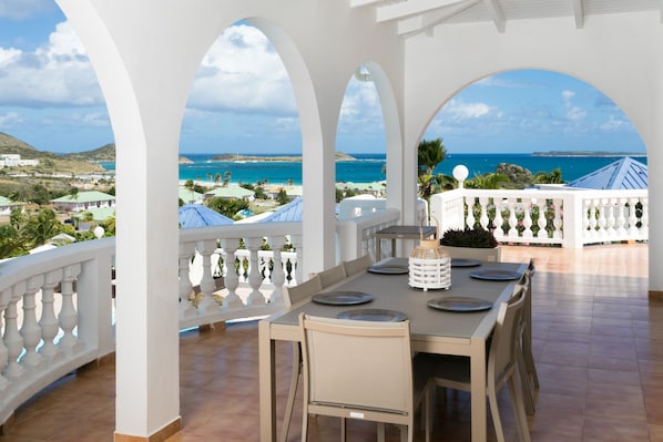 A terrace with a table set for dining, overlooking a coastal landscape with clear skies.