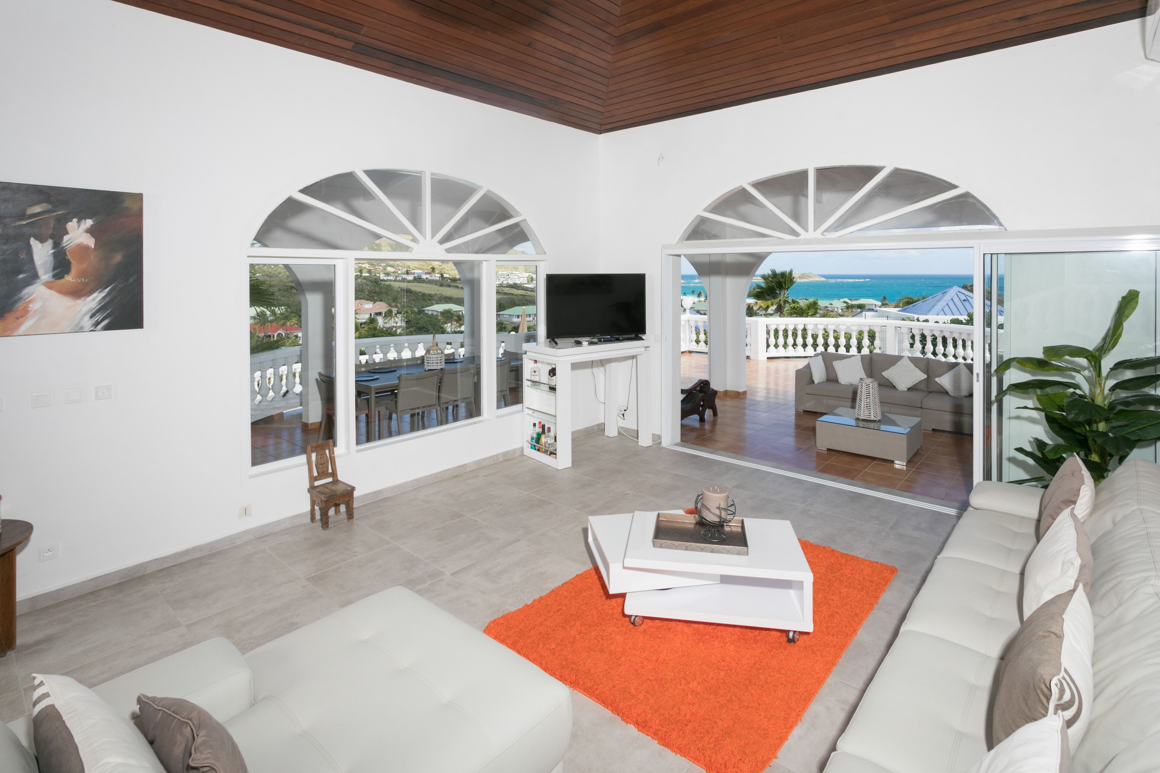 Un vaste espace de vie comprenant un grand canapé, une table basse et un téléviseur. Deux portes en arc donnent accès à un balcon offrant une vue sur la mer.