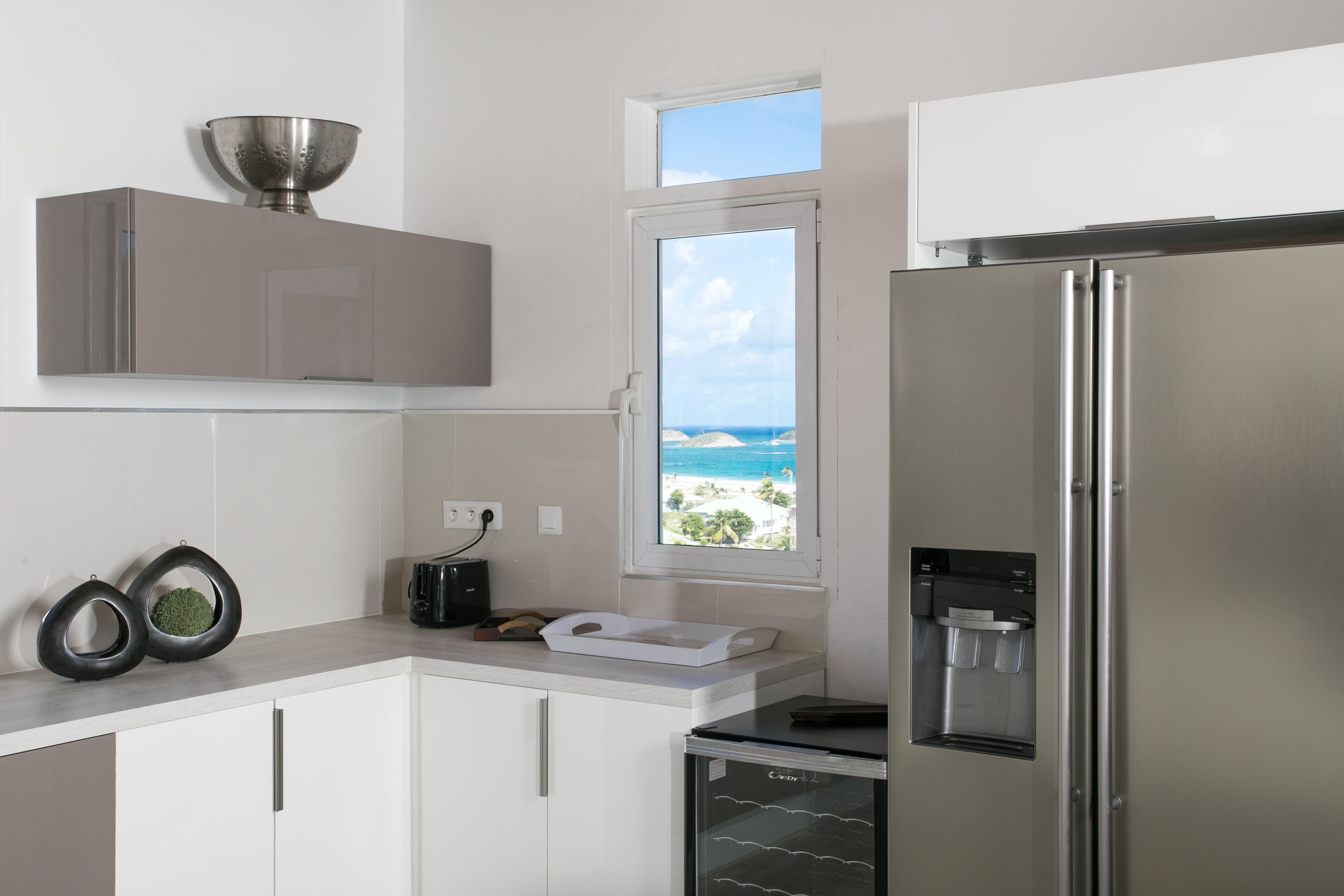 A modern kitchen with a stainless steel refrigerator, a water dispenser, and a view of the ocean through a window.