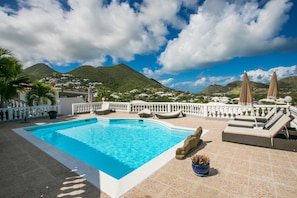 A swimming pool with lounge chairs and umbrellas, surrounded by a white fence and palm trees, with mountains in the background.