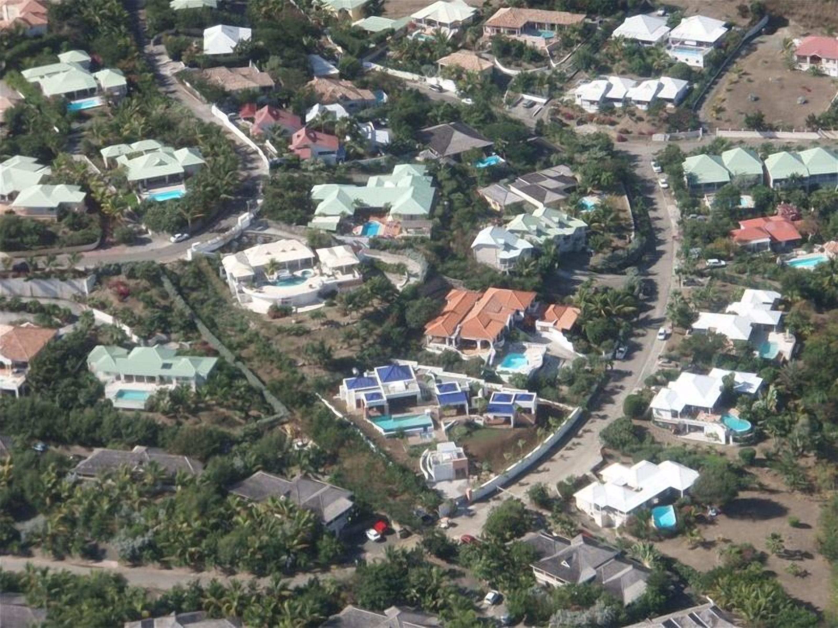 An aerial view of a residential area with multiple houses, some with swimming pools, surrounded by greenery.