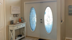Een gang met een witte deur met glas-in-loodramen, een witte bijzettafel met decoratieve voorwerpen en een hanglamp.
