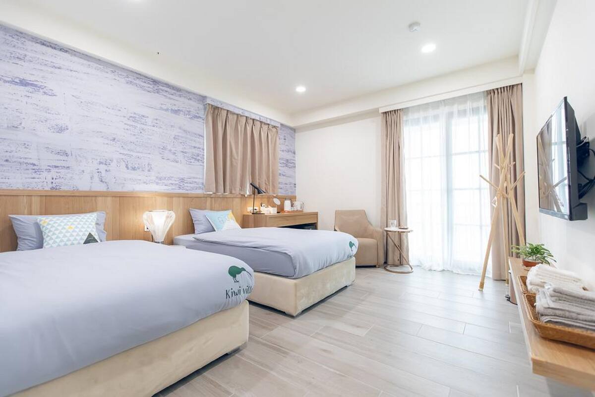 A hotel room with two beds, a TV, a desk, a chair, a window with curtains, and a wall-mounted TV.