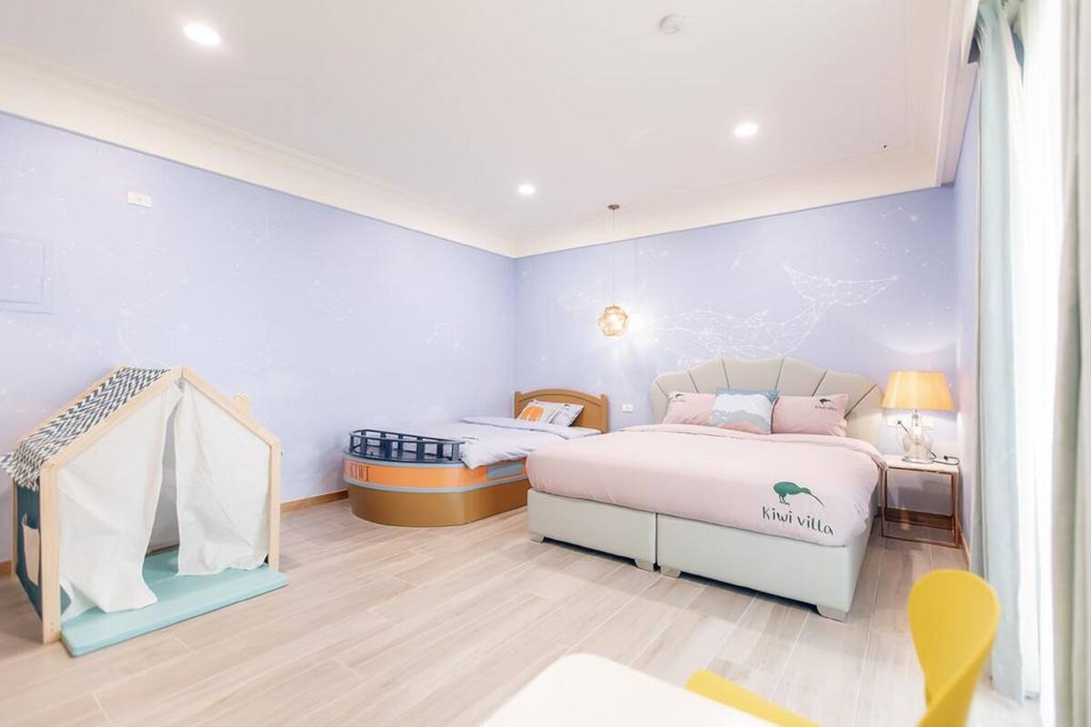 A bedroom with a bed, a small playhouse, a bedside table with a lamp, and a window with curtains.