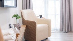 A modern room with a large window, a flat-screen TV mounted on the wall, a beige armchair, a small table with a plant, and neatly folded towels on a shelf. - Kiwi Villa (Magong)