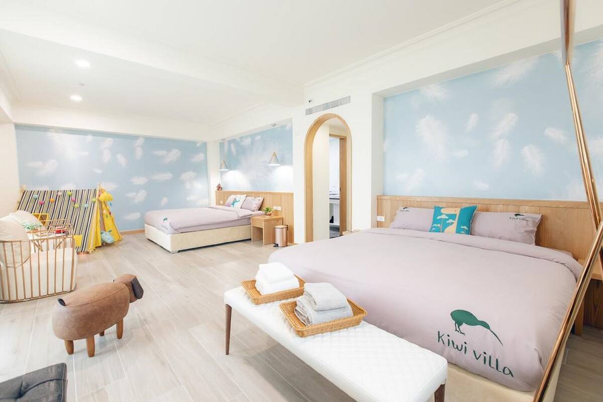 A hotel room with a large bed, a smaller bed, a bench with towels, a baby playpen, and a wall mural.