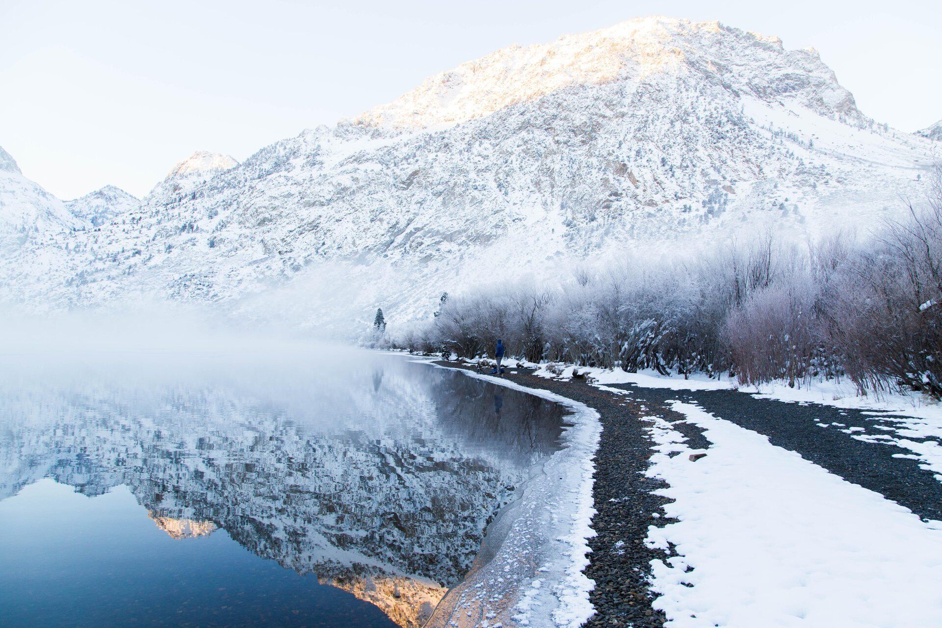 A serene winter landscape with a snow-covered mountain, a calm river reflecting the scenery, and a person walking along the riverbank.